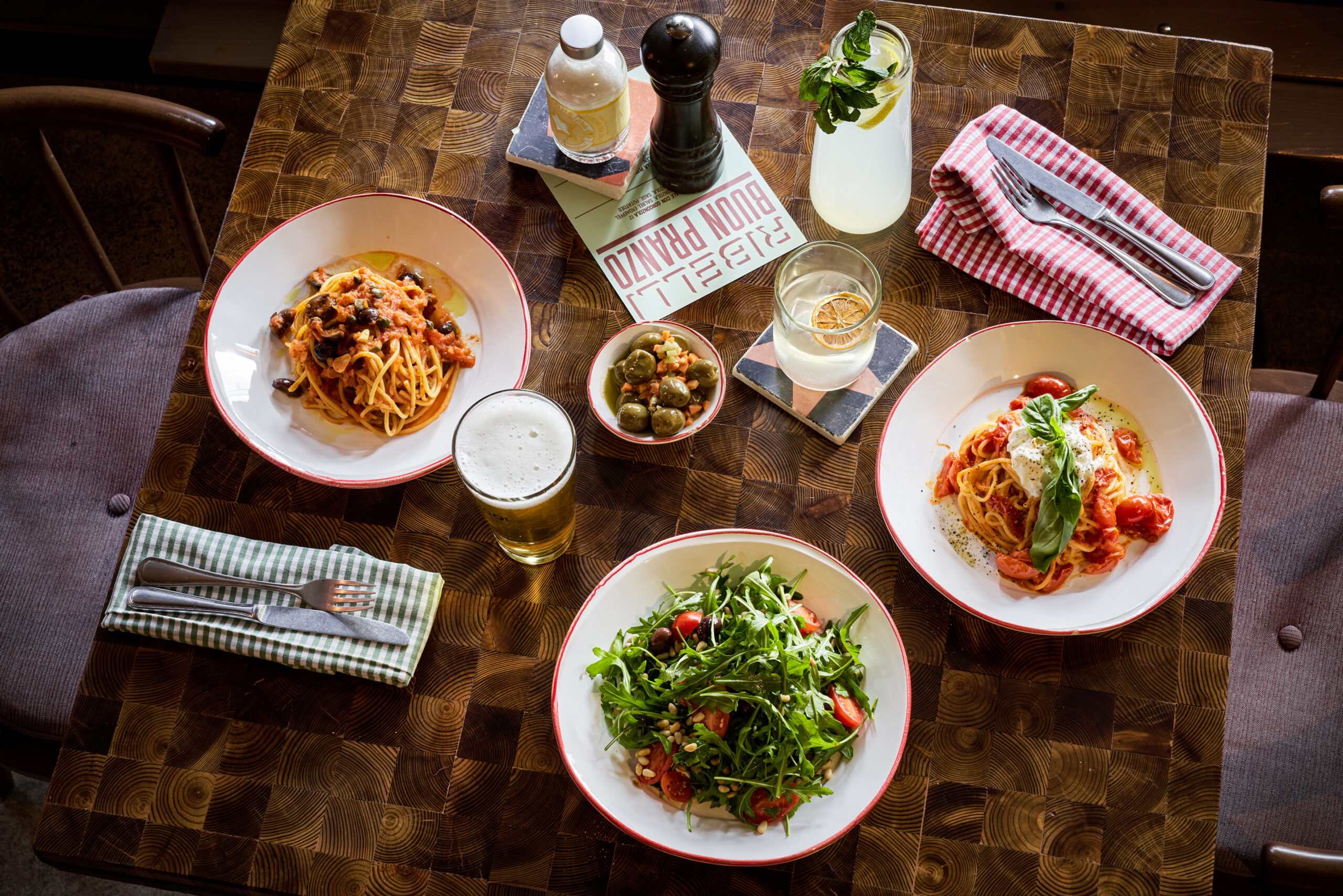 A wooden table with several Italian dishes: pasta, a salad, and olives. Lemonade and beer is next to the plates