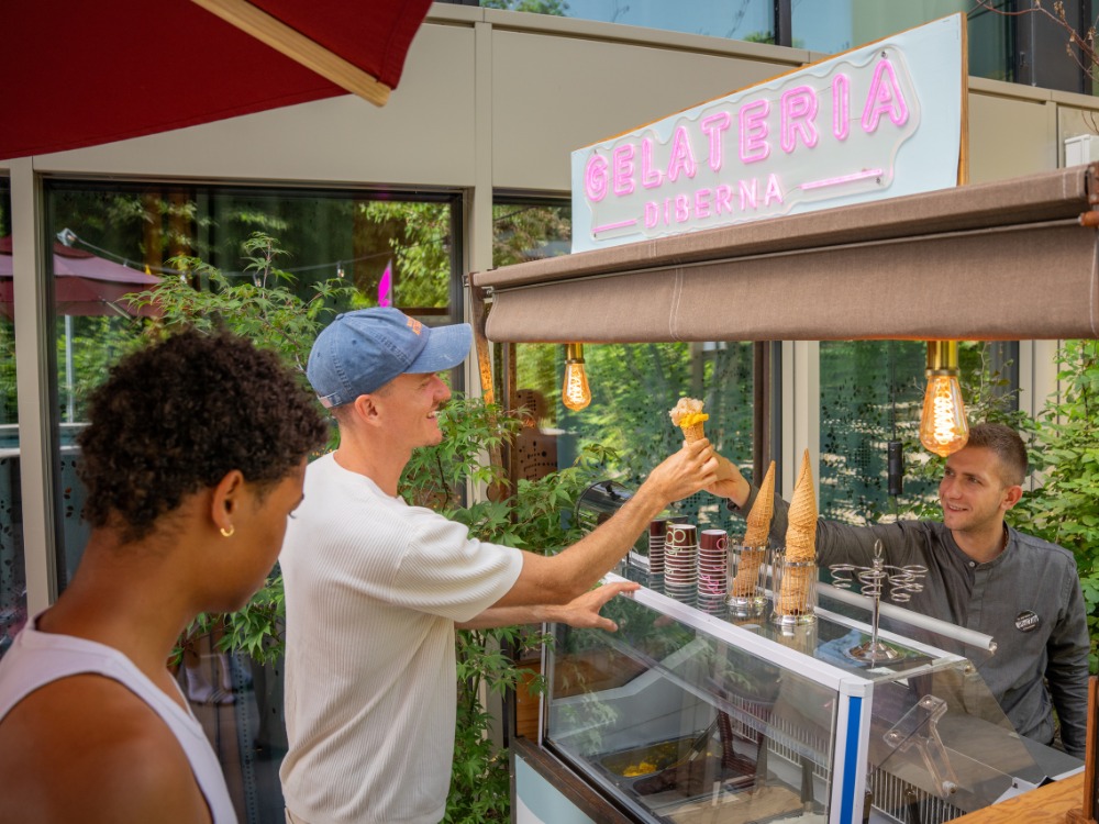 A man in a blue cap smiles while receiving an ice cream from a vendor at "Gelateria di Berna." Another person waits; the mood is cheerful.