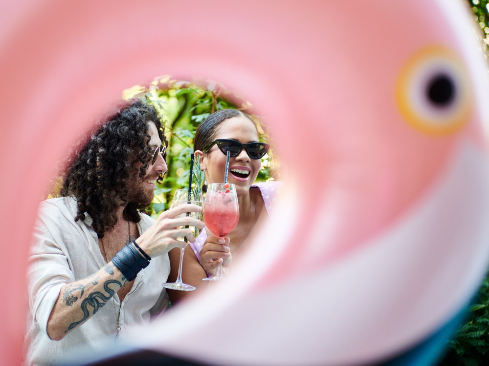 Two people smiling and toasting with cocktails, framed by a pink inflatable flamingo. They are outdoors, conveying a fun, summery vibe.