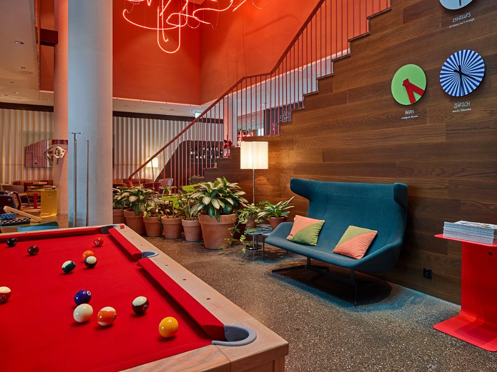 Eine lebhafte Lounge mit einem roten Billardtisch, einem blaugrünen Stuhl, Topfpflanzen und einer leuchtend orangefarbenen Leuchte schafft eine gemütliche, verspielte Atmosphäre.