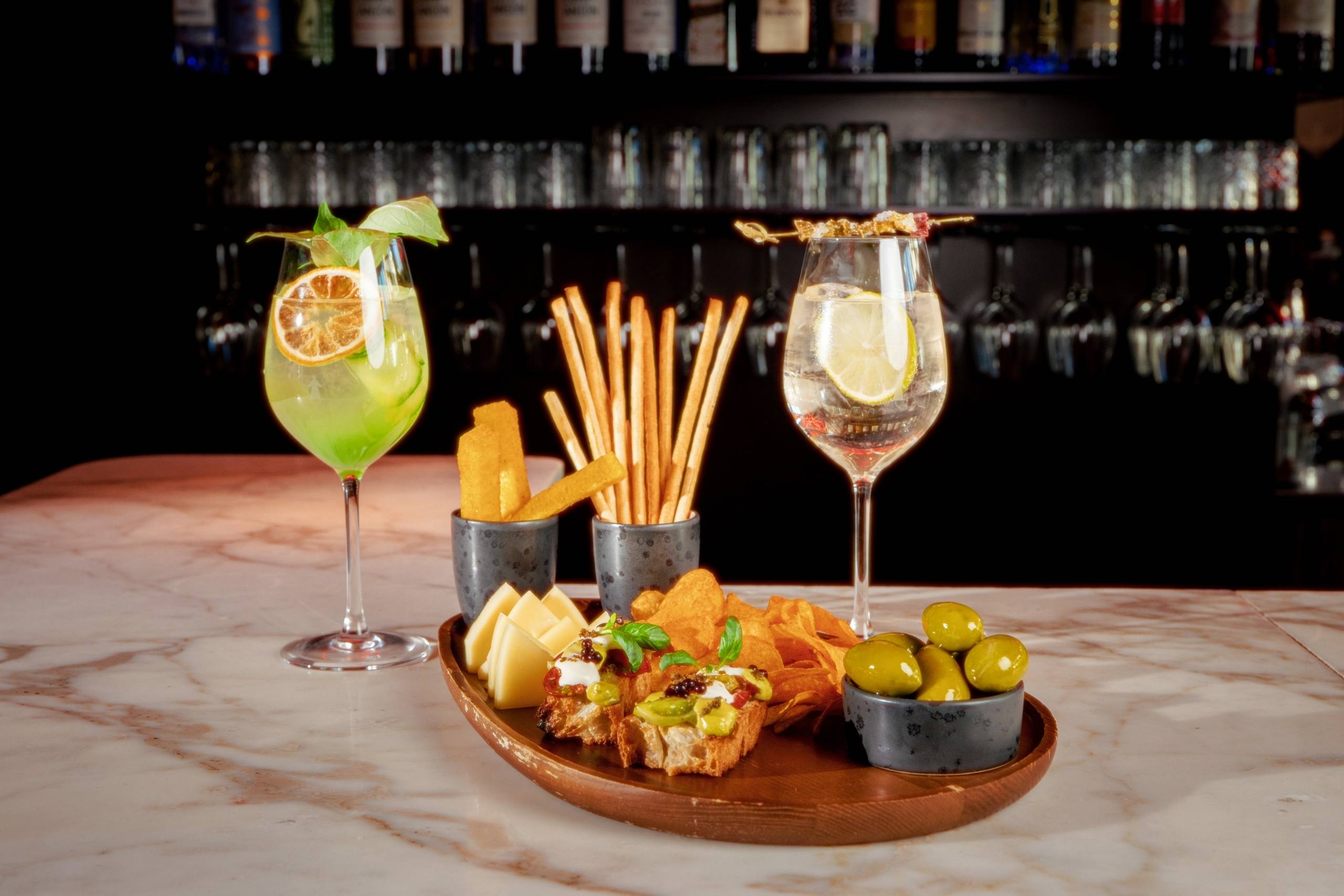 Eine stilvolle Bar-Anrichtung mit zwei Cocktails und Garnituren, umgeben von einer Platte mit Käse, Grissini, Chips und Oliven auf einer Marmor-Theke.