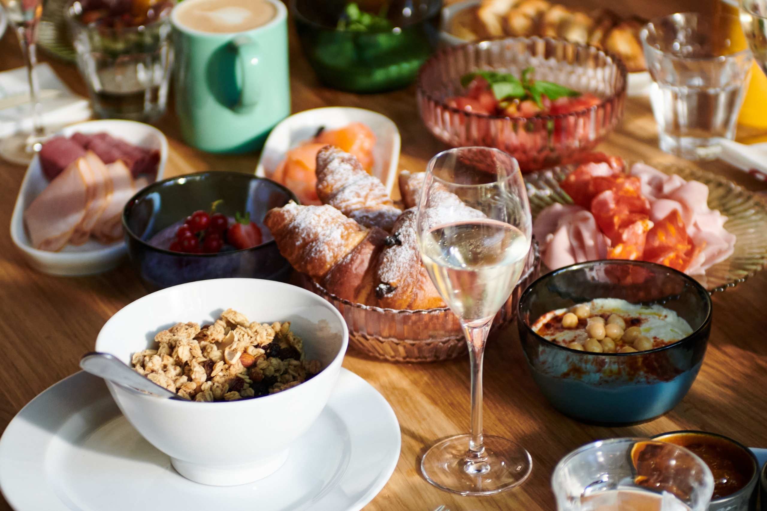 Auf einem Holztisch steht ein reichhaltiges Frühstücksbuffet bereit, darunter eine Schüssel Müsli, Croissants, Aufschnitt, frisches Obst und ein Glas Champagner, was eine einladende, genussvolle Atmosphäre schafft.