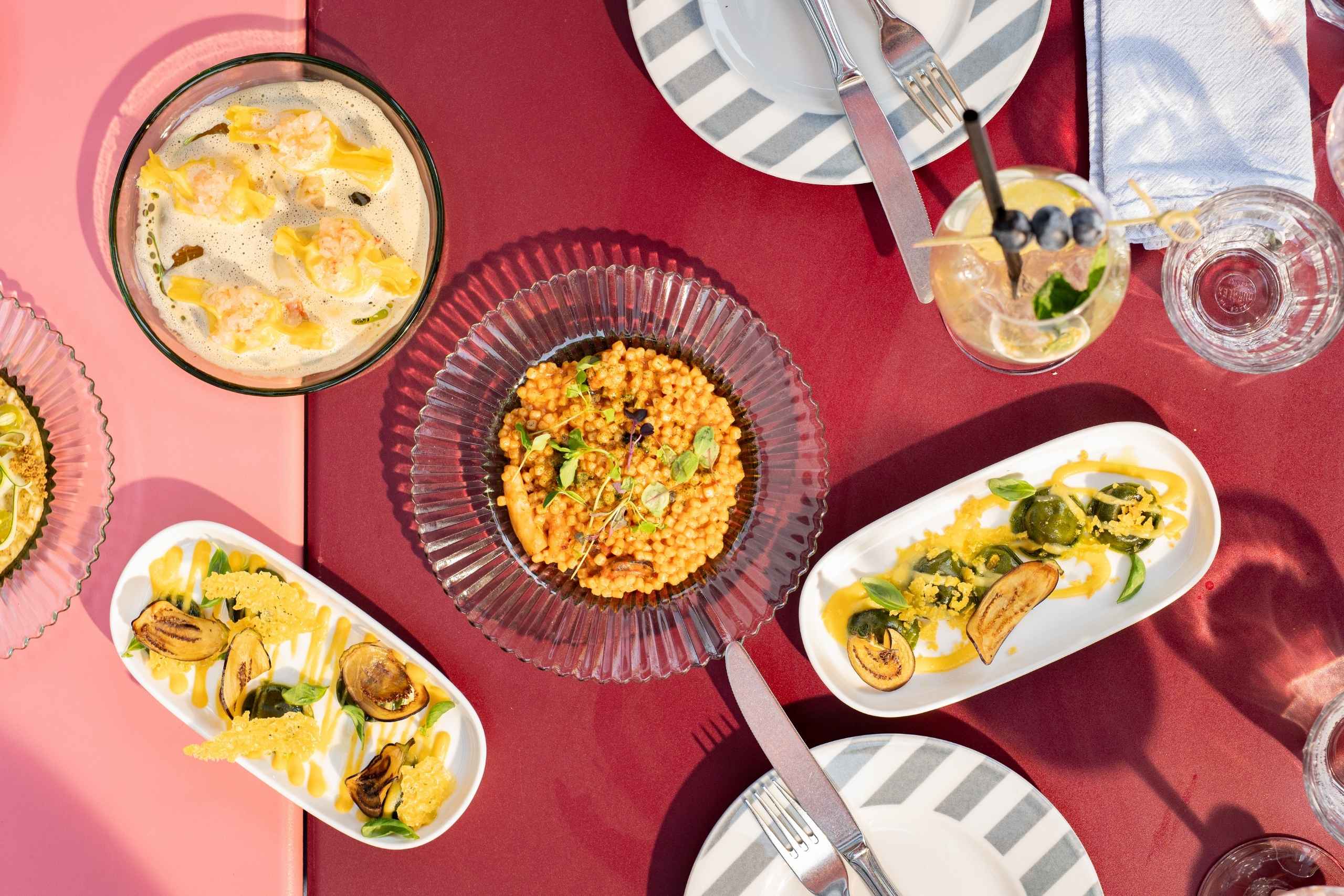 Aerial view of a colorful table setting with diverse dishes, including dumplings in broth, couscous, and garnished vegetables. Plates, utensils, and a drink are also present, suggesting a vibrant culinary experience.
