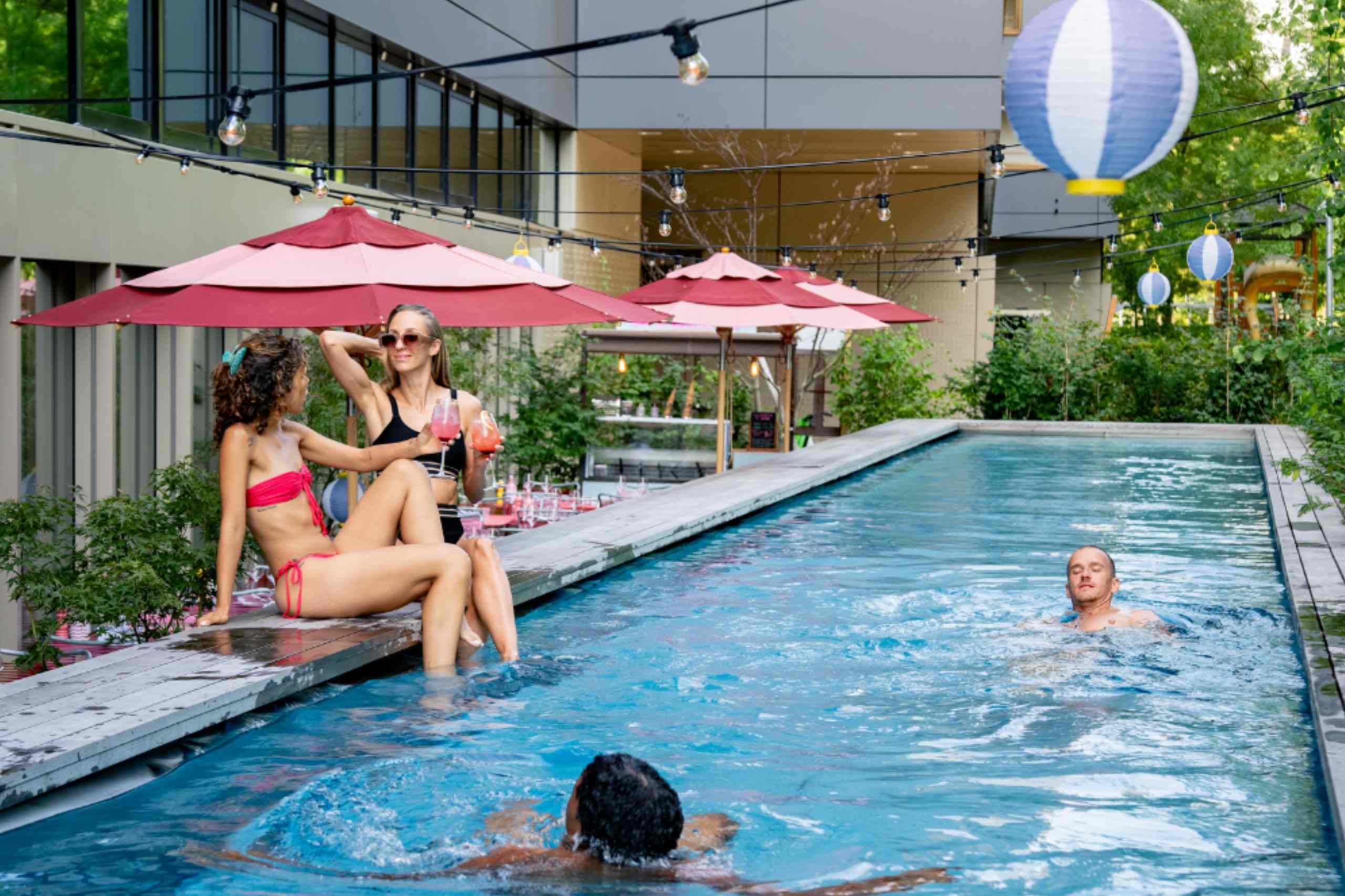 Menschen, die sich in einem rechteckigen Außenpool mit Lichterketten und roten Sonnenschirmen in modernem Ambiente entspannen und schwimmen.