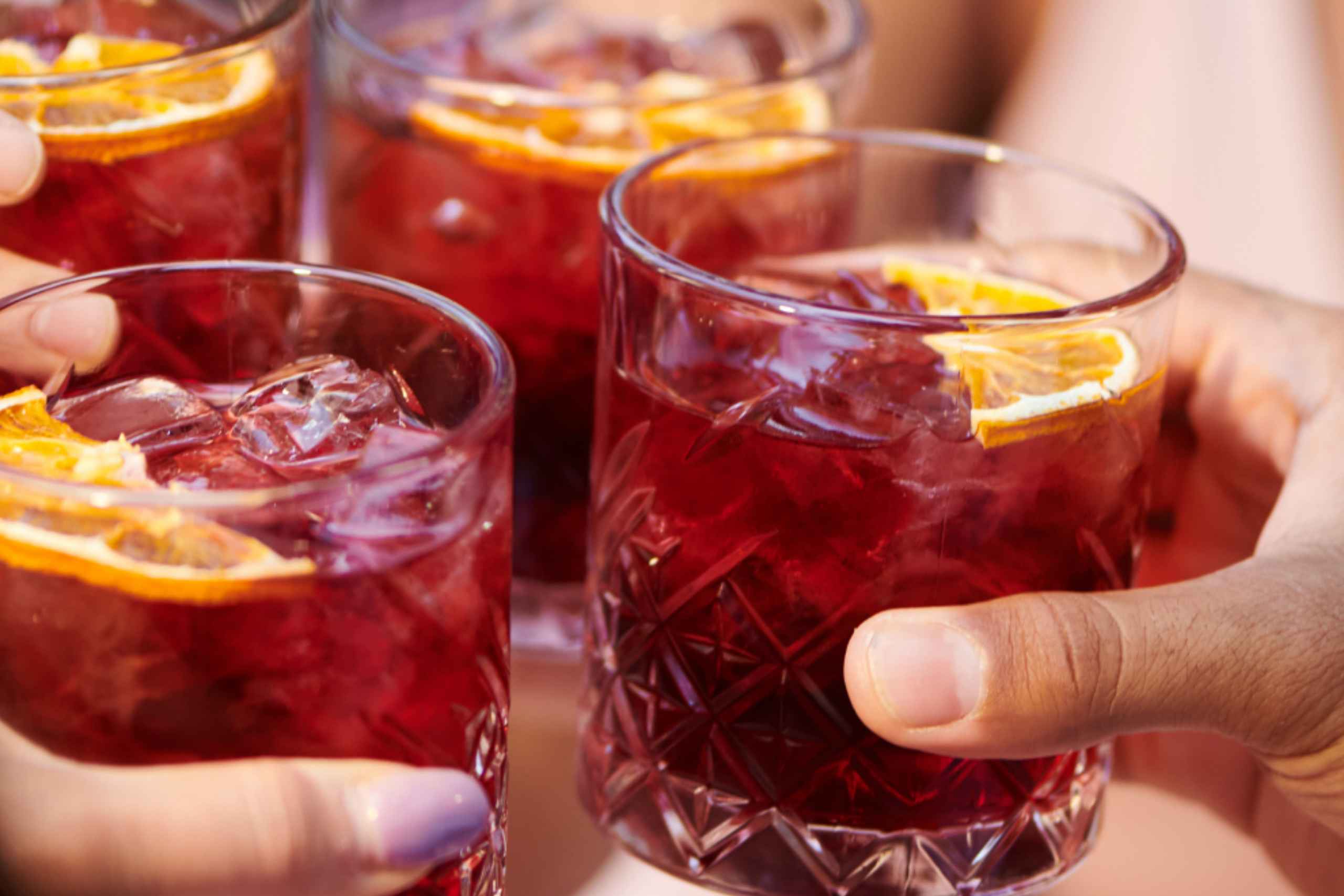 Nahaufnahme von Händen, die Kristallgläser halten und aneinander stoßen, die mit einem leuchtend roten Cocktail, Eiswürfeln und getrockneten Orangenscheiben als Garnitur gefüllt sind, während einer sonnigen Veranstaltung im Freien.